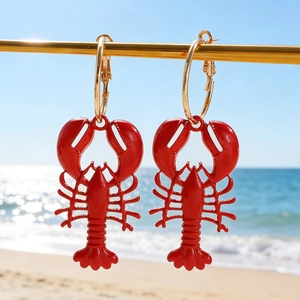 Vibrant red lobster charm earrings hanging on gold hoops with a sunny beach background