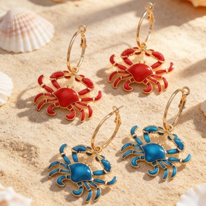 Close-up of red and blue enamel crab earrings on a beach with natural shells