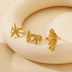 Three gold starfish and shell open rings set on a textured beige fabric background