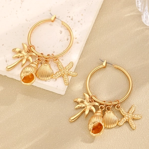 Detail of gold starfish and seashell charms on huggie hoop earrings over a sandy background