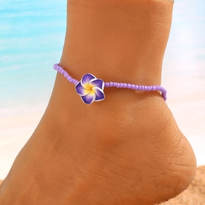 Detalle de una tobillera de cuentas moradas con una flor de plumeria en fondo de playa