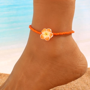 Detalle de tobillera bohemia con cuentas naranjas y colgante de flor de plumeria en playa