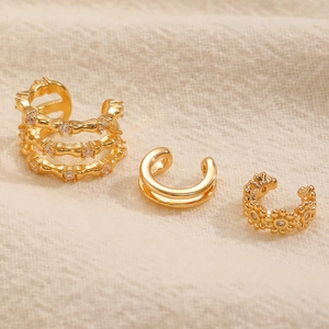 Dettaglio di tre ear cuff dorati a forma di C con zirconi su sfondo di tessuto testurizzato