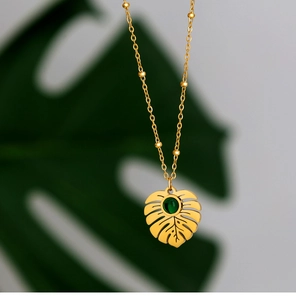 Detail showing a gold monstera leaf pendant with a green gemstone on a beaded chain