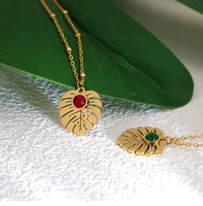 Macro detail of tropical monstera leaf pendants with red and green gems on gold ball chains