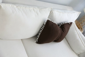 Close-up of a brown striped decorative pillow with white pom-pom edges on a white sofa
