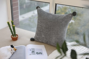 Close-up of gray velvet cushion with plush pompoms on a cozy window sill setting