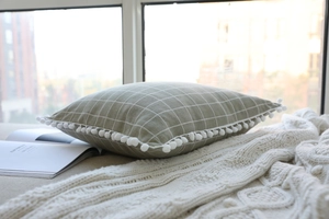 Close-up of sage green checkered pillow with white pom-pom edges on a cozy blanket