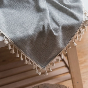 Grey linen textured tablecloth with elegant cream tassels draped over a wooden edge