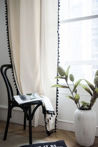 Detail of cream linen curtain with black pompom fringe draping over a wooden chair