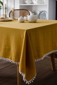 Detail of yellow washed cotton tablecloth with white pom-pom trim on a wooden table