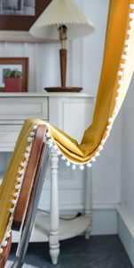 Close-up of bright yellow semi-sheer curtain with white pom-pom trim on a wooden chair