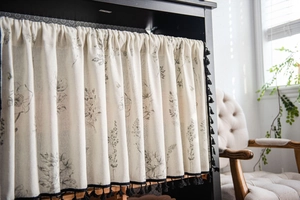 Detail of floral print cafe curtain with black tassel fringe on a rustic cabinet