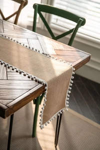 Detail of natural jute table runner with white pom-pom trim on a dark wood table
