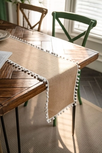 Close-up of natural burlap table runner with white pom-pom trim on wood table