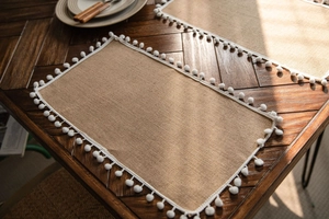 Detail view of natural jute placemat featuring white pom-pom trim on dark wooden table