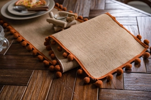Detail of natural burlap table runner with orange pom-pom fringe on a wooden table