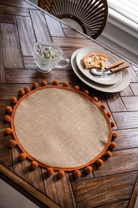 Detailed view of round natural jute placemat with orange pom-pom edge on a wood table