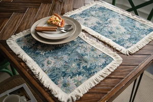 Close-up of blue floral jacquard placemats with ivory fringe on a rustic wooden table