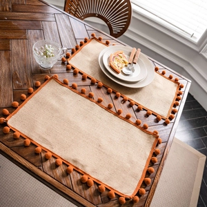 Set of two rectangular jute placemats with orange pom-pom fringe on a wooden table