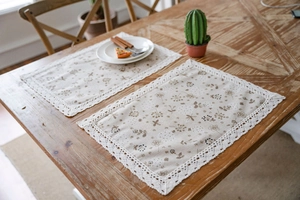 Dettaglio di tovagliette beige con motivi floreali caffè e bordi in pizzo bianco