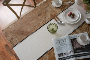 Detail of beige fabric table runner with black pompom fringe on dark wood table