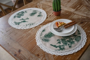 Detail of round cotton linen placemat with pine cone prints and white lace edges on wood table
