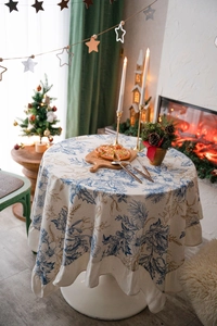 Close-up of beige round tablecloth featuring pine cones, holly patterns and ruffled edges