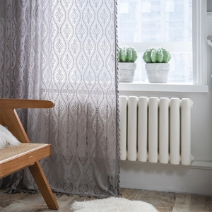 Grey lace curtain with geometric floral patterns and fringe tassels in a bright room