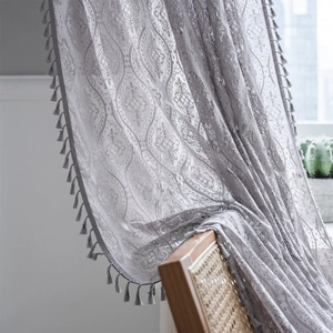 Grey crochet lace curtain with floral patterns and elegant fringe tassels in a rustic room