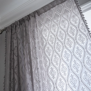 Grey crochet lace curtain with damask patterns and tassel edges in a sunny window
