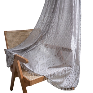 Grey crochet lace curtain with hollow patterns and tassel fringe on a rustic wooden chair