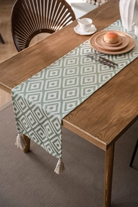 Detail of green geometric jacquard table runner with beige handmade tassels on wood table