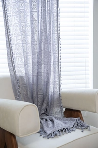 Detail of grey crochet lace curtain with geometric patterns and long tassels on white sofa