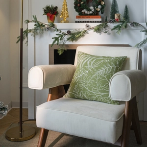 Green Christmas pillow cover with white leaf print on a cozy armchair by the fireplace
