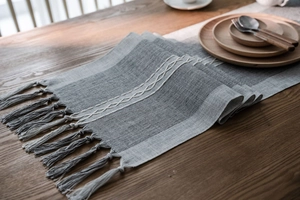Detail of grey jacquard table runner with white embroidery and tassel fringes on wood