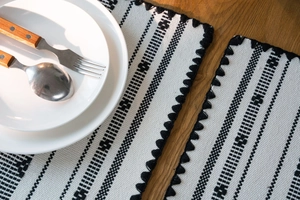 Dettaglio della tovaglietta jacquard geometrica con frange nere su tavolo di legno