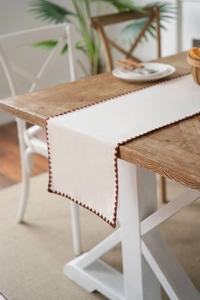 Detail of minimalist cream table runner with brown tassel trim on dark wood table
