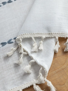 Detailed close-up of white linen fabric with grey geometric embroidery and fringe tassels