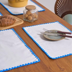 Modern white rectangular dining placemat with blue pom-pom lace trim on a wooden table