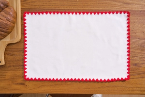 White cotton linen placemat with red crochet edge on dark wooden table with bread tray