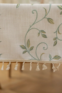Close-up of beige cotton linen tablecloth featuring green leaf embroidery and fringe