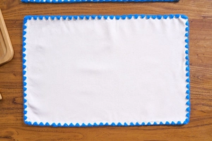 Rectangle white dining placemat with blue pompom trim edge on dark wooden table