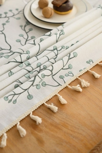 Detailed embroidery of green leaves on an ivory cotton linen table runner with tassels