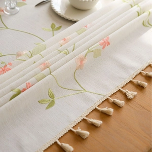 Beige floral embroidered table runner with decorative tassels on a wooden surface