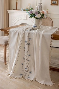 Detail of cream tablecloth with blue floral embroidery and fringe tassels on wood table