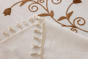 Detailed macro view of beige linen tablecloth featuring brown floral embroidery and fringe