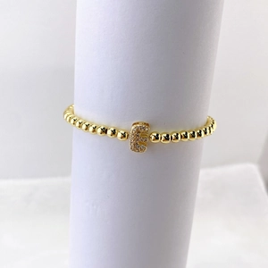 Detalle de pulsera de cuentas doradas con dije de letra E con micropavé sobre fondo blanco