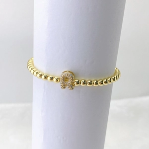 Detalle de pulsera de cuentas doradas con dije de letra R en circonitas sobre fondo blanco