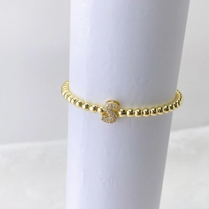 Detalle de pulsera de cuentas doradas con un dije de letra S de cristal en micro pavé